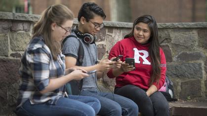 three students sit on a bench browsing their phones
